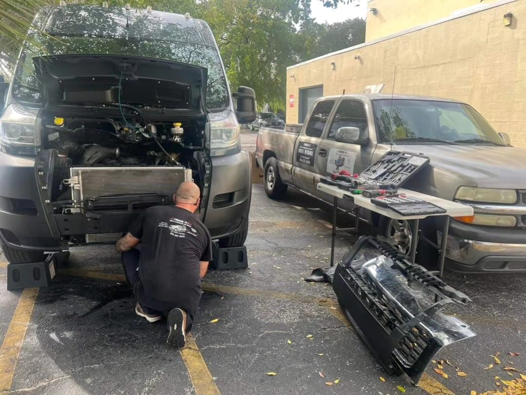Person working on engine of van in parking lot with two service trucks and equipment visible